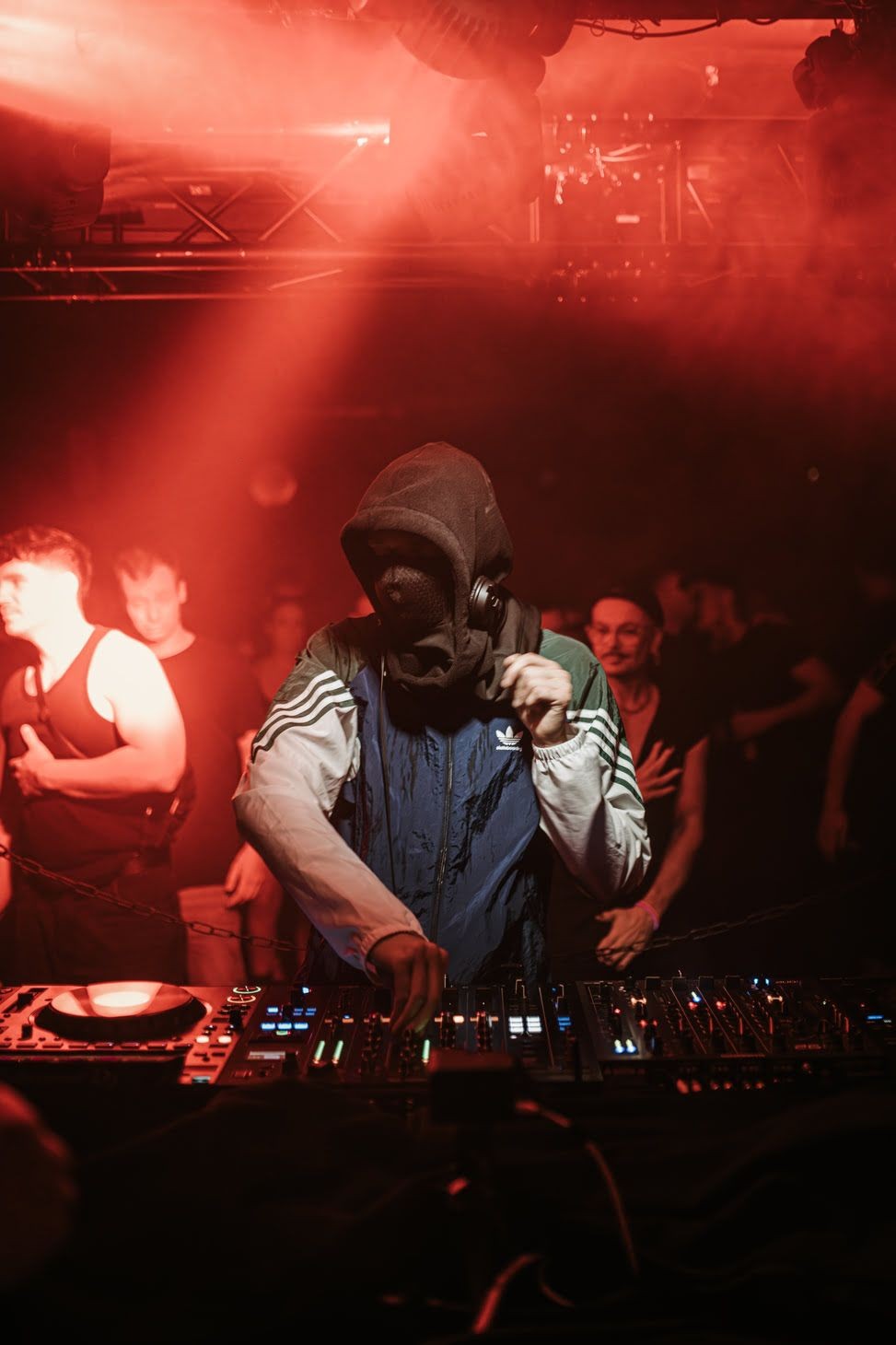 DJ wearing a hoodie and headphones performing on stage with turntables and mixing equipment under red lights.