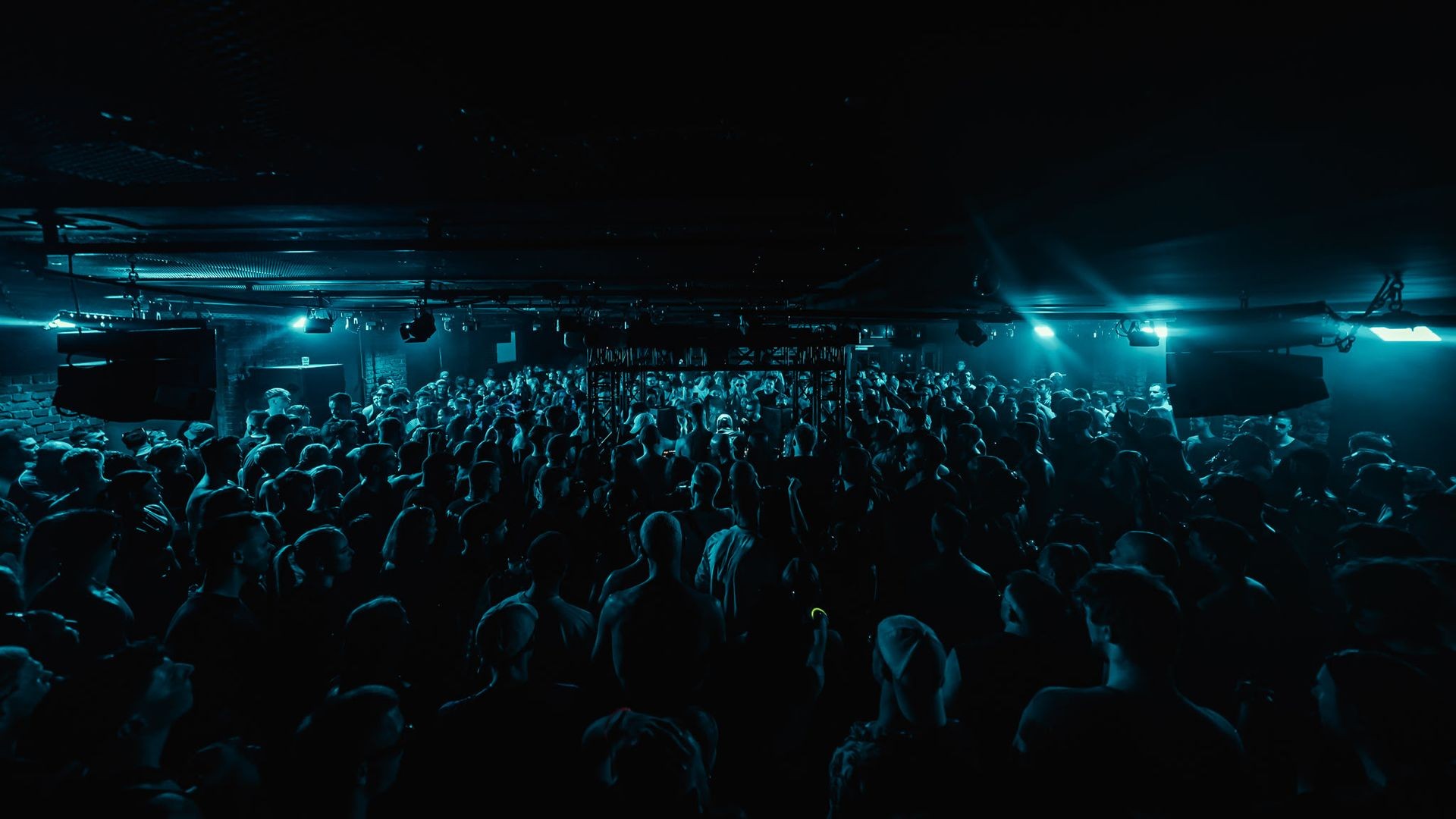 Crowd of people in a club with teal lighting and music equipment visible on stage.