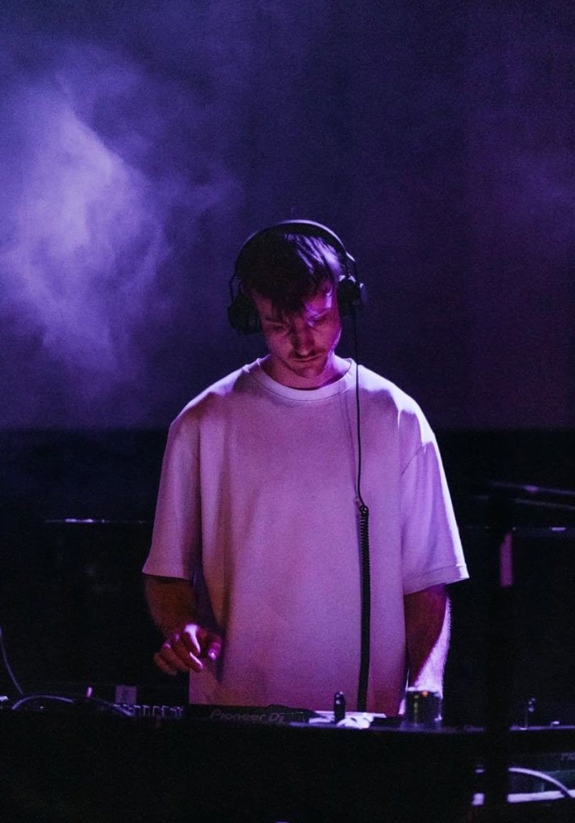 DJ wearing headphones mixing music on a dark stage with purple lighting and smoke effects.