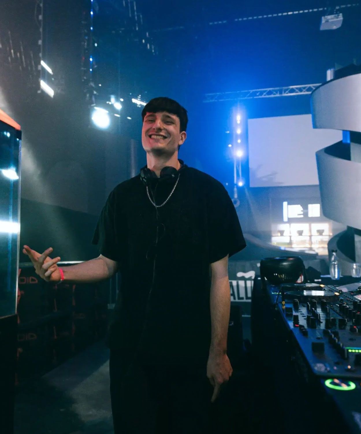 DJ smiling on a nightclub stage with turntables and bright stage lights in the background.