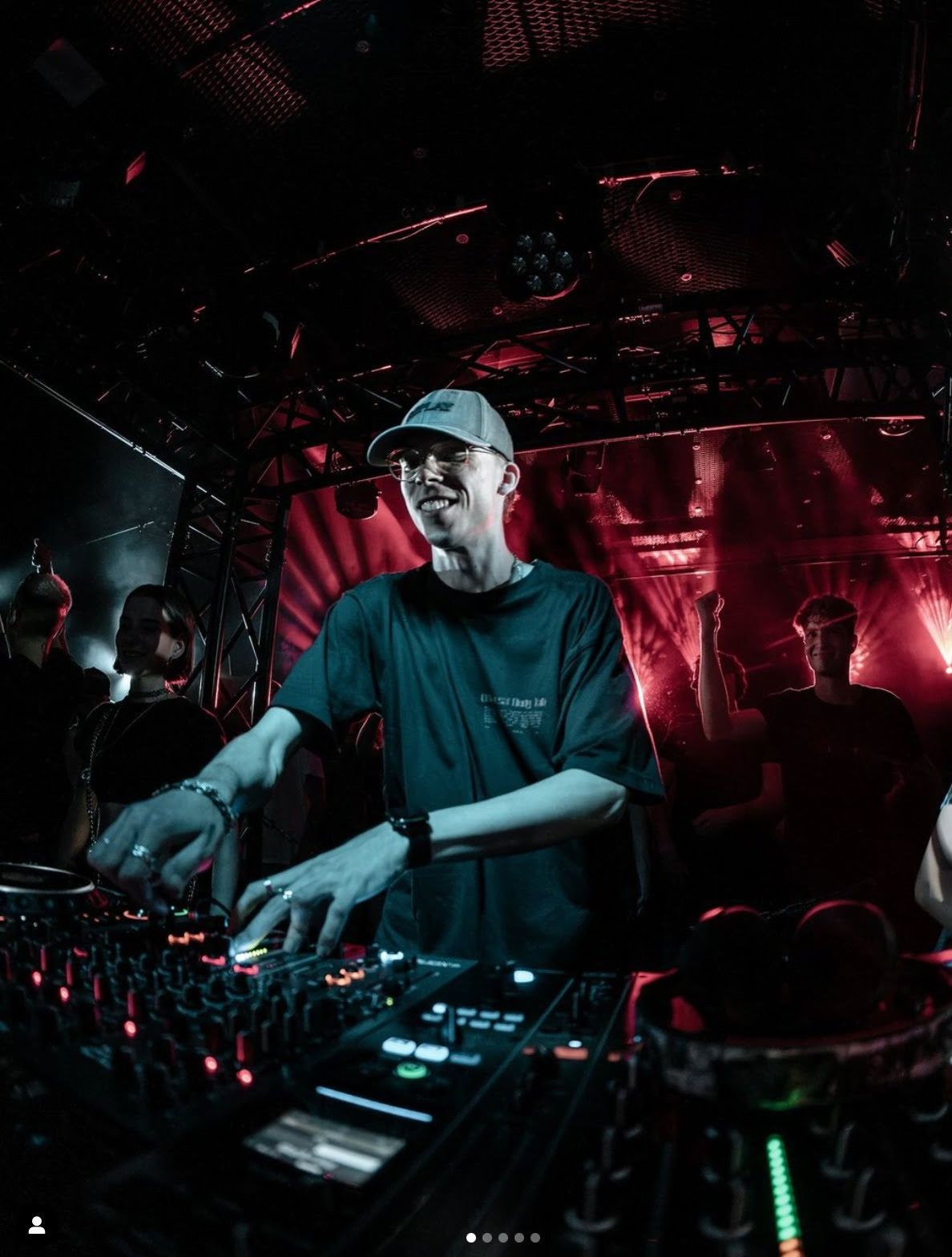 DJ wearing a cap performing in a dimly lit club with red lighting and an enthusiastic crowd.